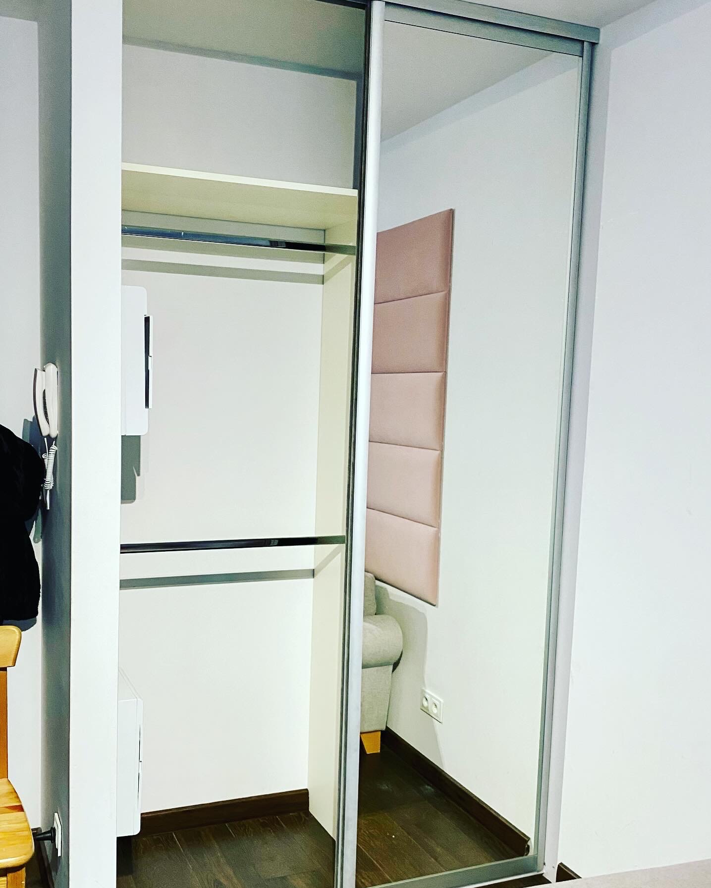 Wnętrze szafy przesuwnej z drążkami i półką, z widocznym odbiciem fragmentu pokoju: pudrowy zagłówek i narożnik.