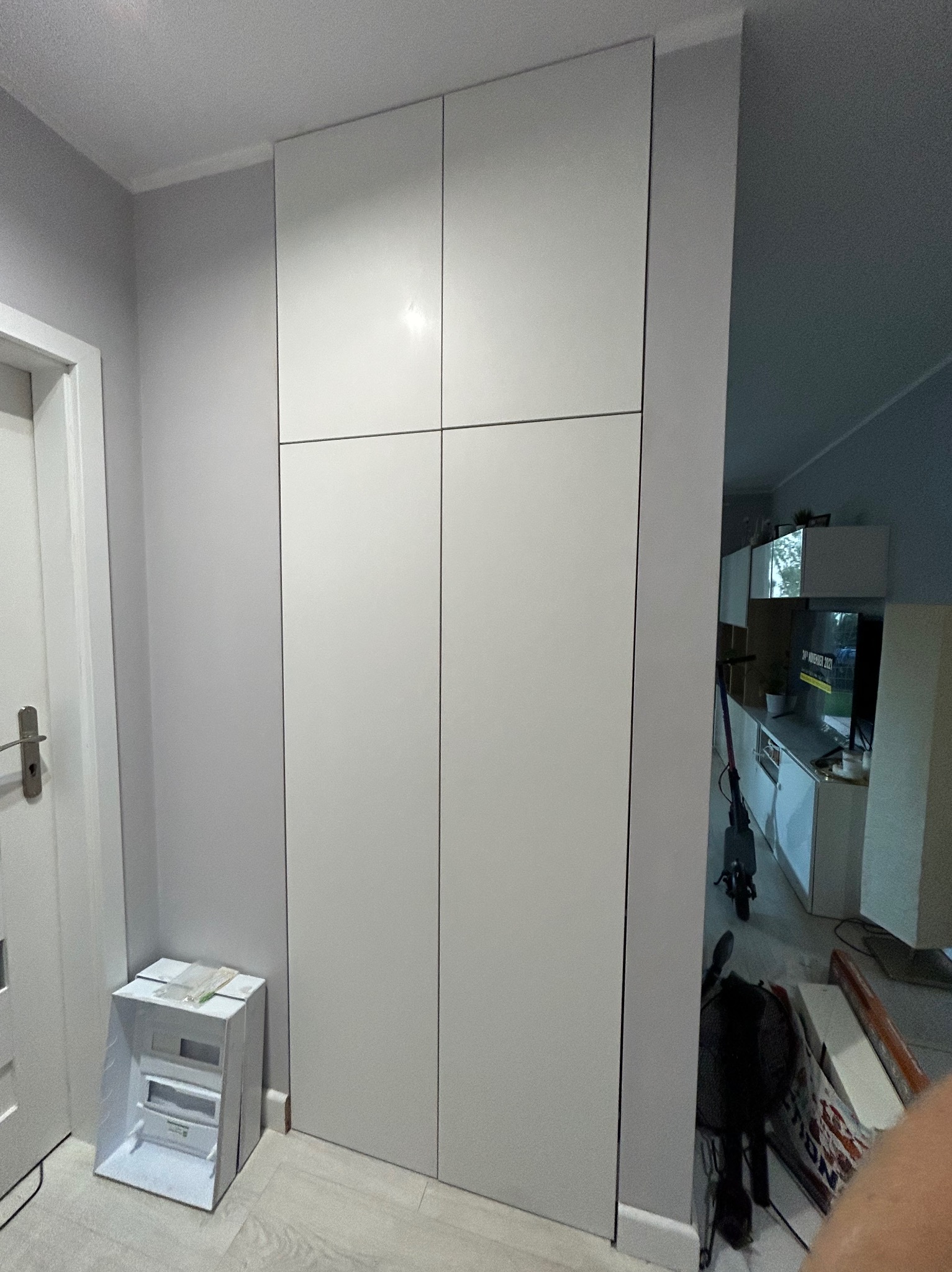 Biała, czterodrzwiowa szafa wnękowa z matowymi frontami, dopasowana do wysokości pomieszczenia, z widocznym fragmentem drzwi wejściowych po lewej stronie i wnętrzem pokoju po prawej.