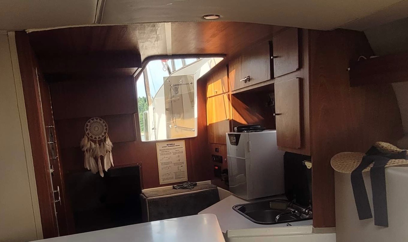 Wnętrze kabiny jachtu z drewnianą zabudową, widoczne szafki, zlewozmywak, mała lodówka, okno z widokiem na zewnątrz i łapacz snów na ścianie.