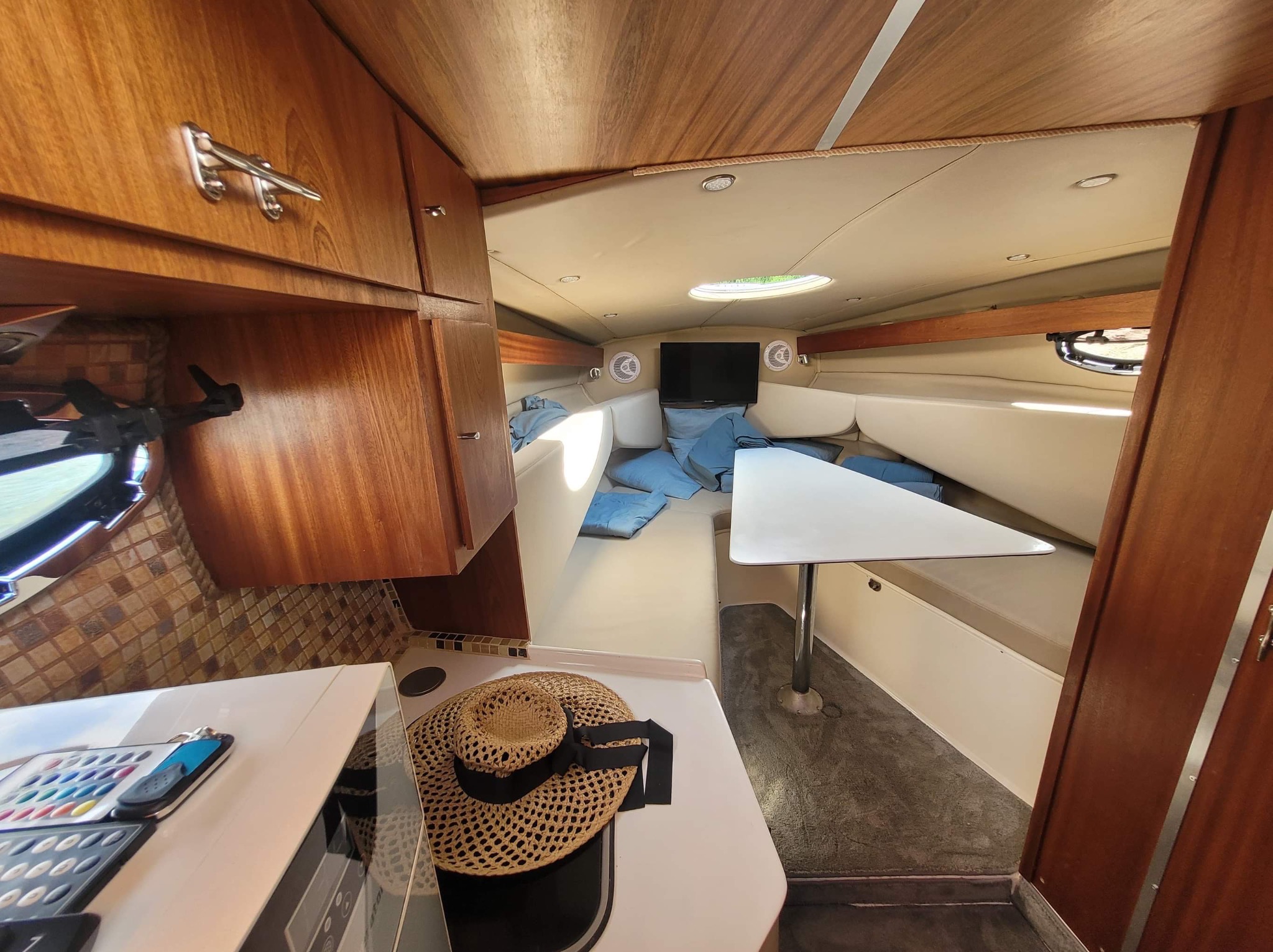 Wnętrze jachtu z drewnianymi szafkami, białą tapicerką, małym stolikiem i telewizorem. Na blacie kuchennym leży słomkowy kapelusz.