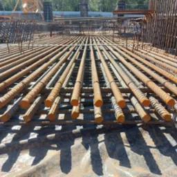 Siatka zbrojeniowa ułożona na folii budowlanej, widoczne pręty zbrojeniowe z wyraźną fakturą i śladami korozji, w tle elementy konstrukcji żelbetowej i żuraw budowlany.