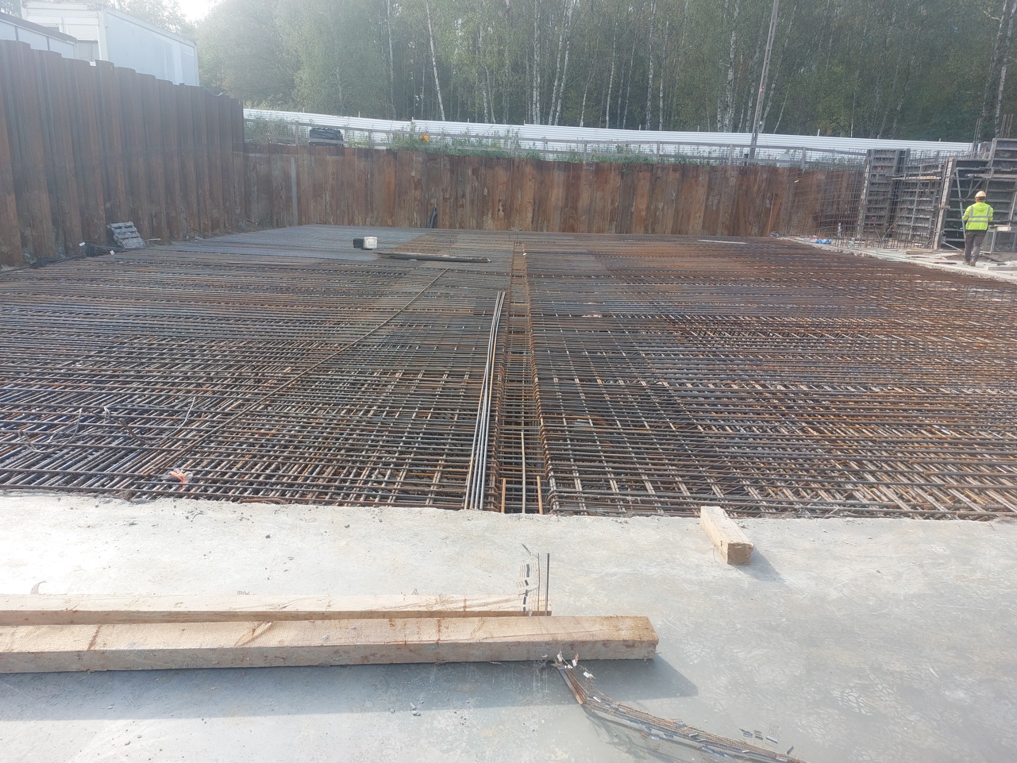 Siatka zbrojeniowa na betonowej płycie fundamentowej z deskami szalunkowymi w pierwszym planie, widoczna osoba w odblaskowej kamizelce w tle.