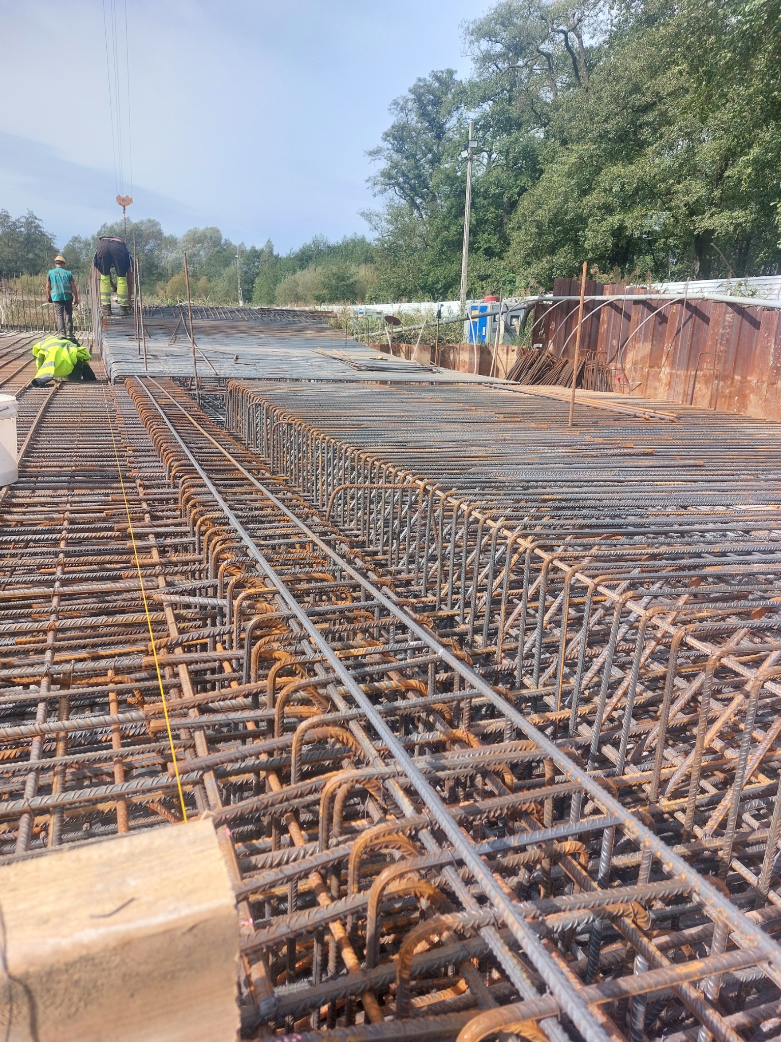 Konstrukcja zbrojeniowa z widocznymi pracownikami w oddali, przygotowana do zalania betonem; widoczne liczne pręty zbrojeniowe i szalunki.