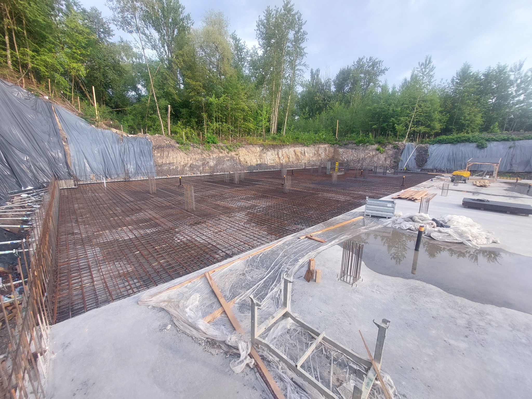 Szeroki kadr na przygotowany teren pod fundamenty z widoczną siatką zbrojeniową i wystającymi prętami, częściowo zalany wodą, z zabezpieczonym skarpą w tle.