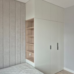 WOOD MEBLE SPÓŁKA Z OGRANICZONĄ ODPOWIEDZIALNOŚCIĄ - Jasnoszara szafa wnękowa z otwartymi półkami z jasnego drewna i czarnymi uchwytami, przylegająca do tapicerowanego panelu ściennego nad łóżkiem.