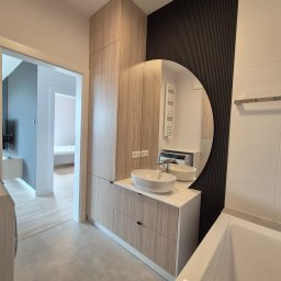 WOOD MEBLE SPÓŁKA Z OGRANICZONĄ ODPOWIEDZIALNOŚCIĄ - Nowoczesna łazienka z szafką na wymiar z drewnopodobnym frontem, owalną umywalką nablatową i okrągłym lustrem na tle ściany z ciemnymi lamelami.