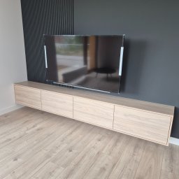 WOOD MEBLE SPÓŁKA Z OGRANICZONĄ ODPOWIEDZIALNOŚCIĄ - Nowoczesna szafka RTV z forniru drewnopodobnego, wisząca pod telewizorem na czarnej ścianie z dekoracyjnymi lamelami.
