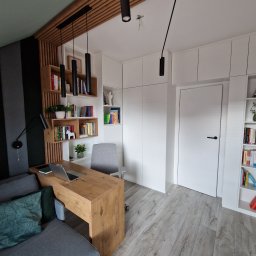 WOOD MEBLE SPÓŁKA Z OGRANICZONĄ ODPOWIEDZIALNOŚCIĄ - Nowoczesny pokój z drewnianym biurkiem, laptopem, szarą sofą i zielonymi poduszkami, drewnianymi półkami na książki i białymi szafami w zabudowie, oświetlony wiszącymi lampami.