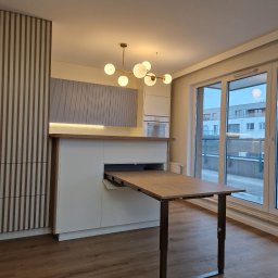 WOOD MEBLE SPÓŁKA Z OGRANICZONĄ ODPOWIEDZIALNOŚCIĄ - Nowoczesna kuchnia z wysuwanym stołem z blatem w kolorze drewna, oświetlenie podszafkowe LED, dekoracyjne panele ścienne z pionowych listew w dwóch kolorach, widok na balkon.