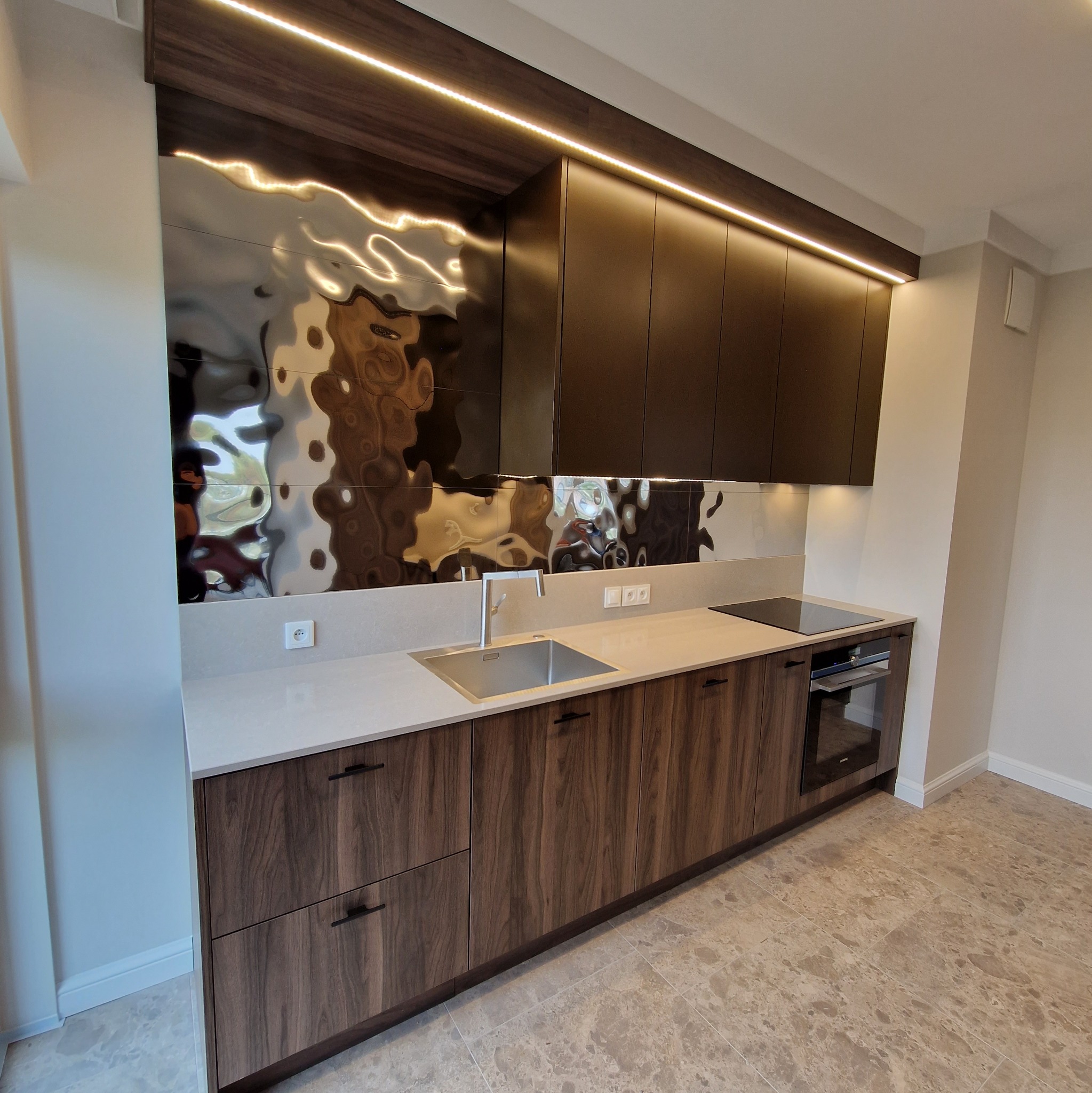 Nowoczesna kuchnia na wymiar z ciemnymi, drewnianymi frontami szafek, jasnym blatem i błyszczącym panelem nad blatem, oświetlona listwą LED