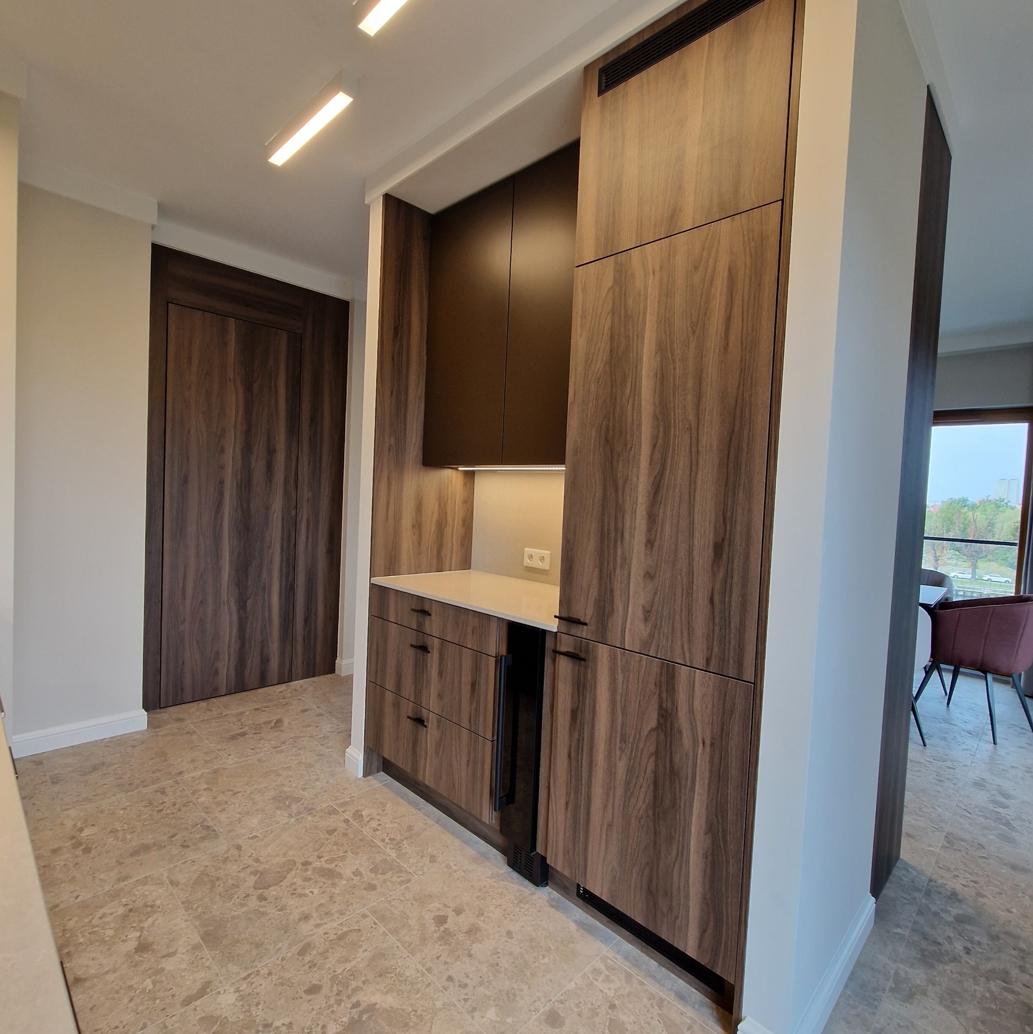 Nowoczesna zabudowa kuchenna z forniru orzechowego, zintegrowana z szafą wnękową o identycznym wykończeniu, oświetlona punktowo pod szafkami wiszącymi, z widocznym fragmentem jadalni w tle.