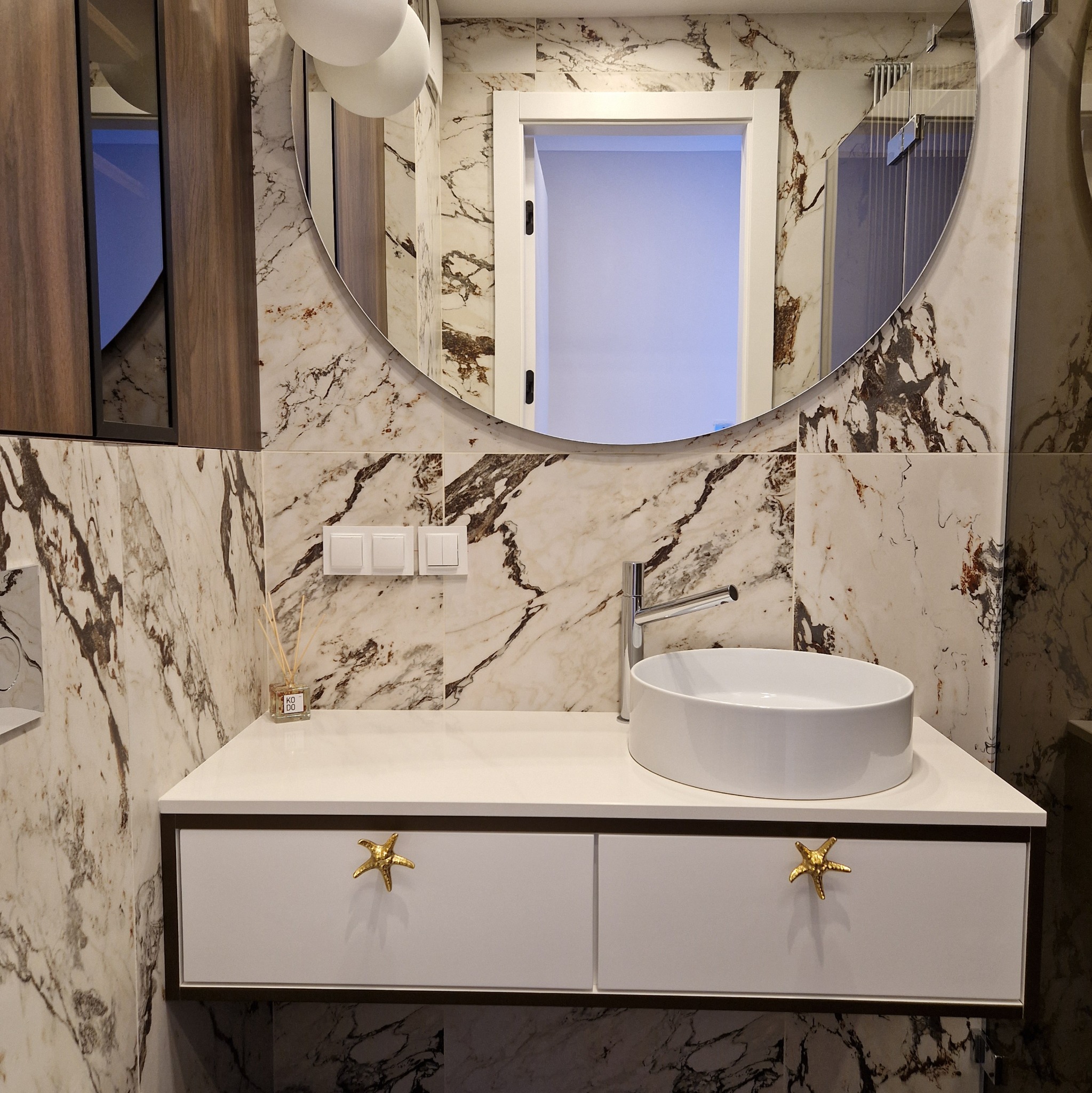 Nowoczesna łazienka z umywalką nablatową, armaturą i szafką z uchwytami w kształcie złotych rozgwiazd na tle marmurowej ściany i okrągłego lustra.