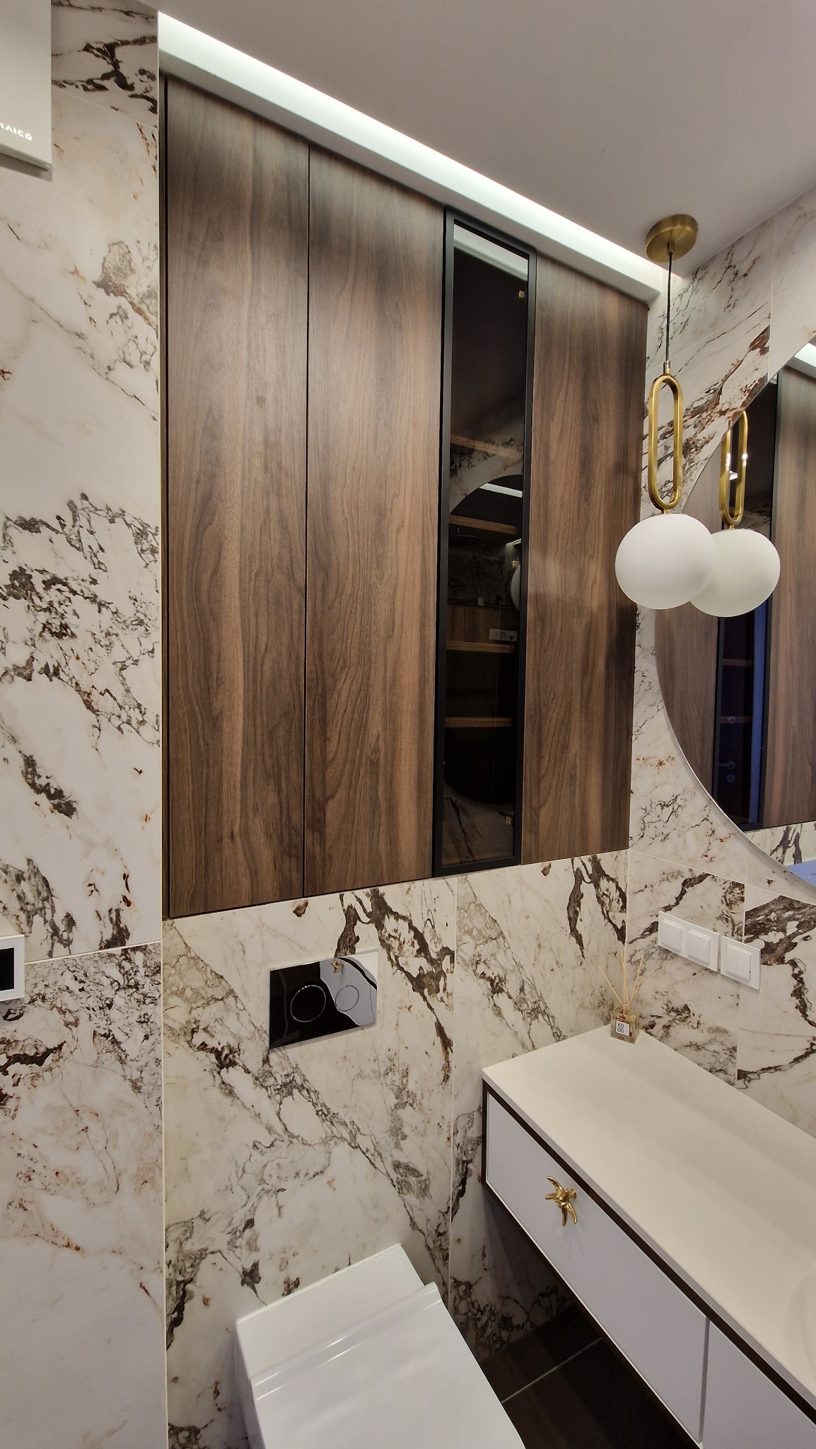 Nowoczesna łazienka z drewnianą szafą wnękową z czarnymi akcentami, marmurowe płytki na ścianach, okrągłe lustro i designerska lampa wisząca z elementami w kolorze złota.