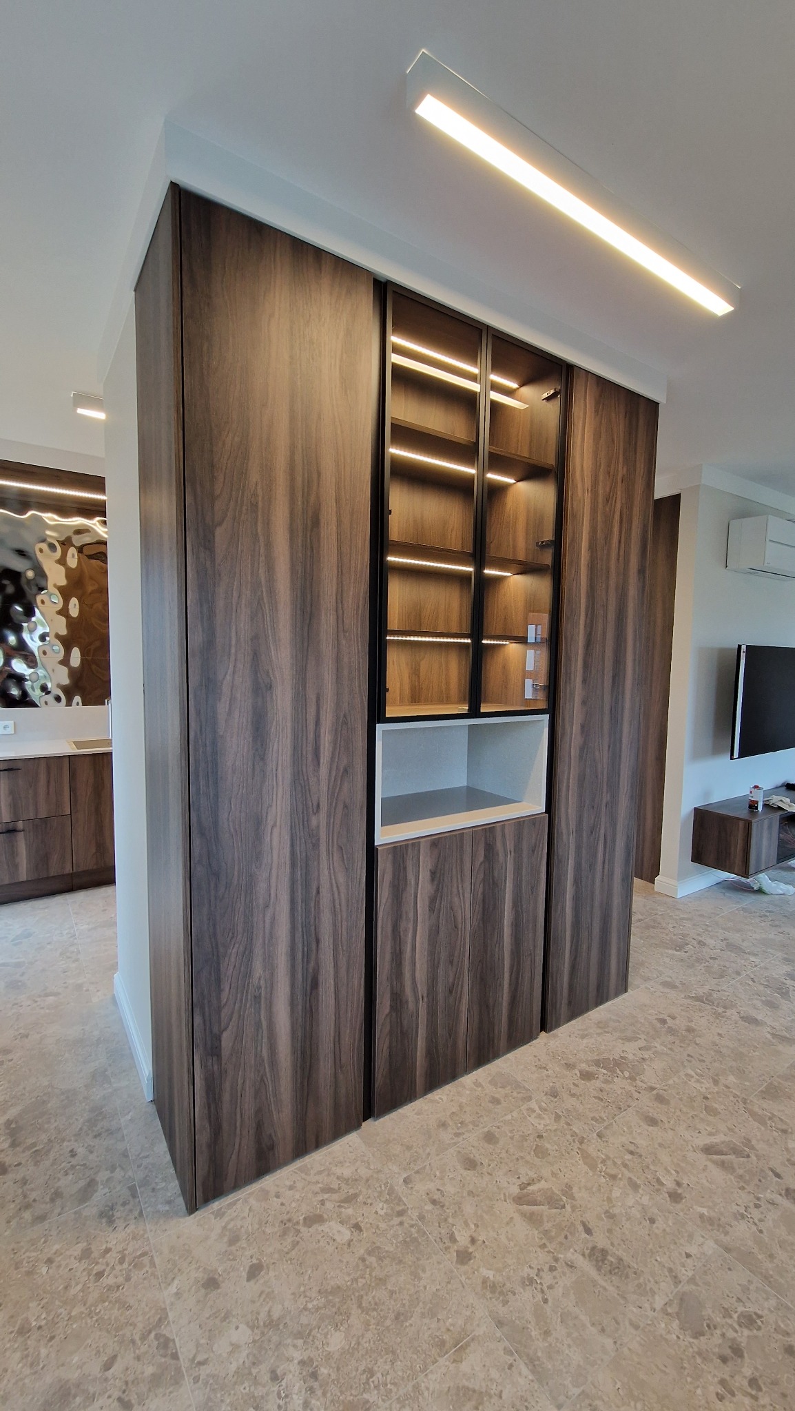 Nowoczesna zabudowa meblowa w salonie z fornirem w odcieniach brązu, podświetlane szklane witryny, biała półka i jasna podłoga z płytek imitujących kamień.