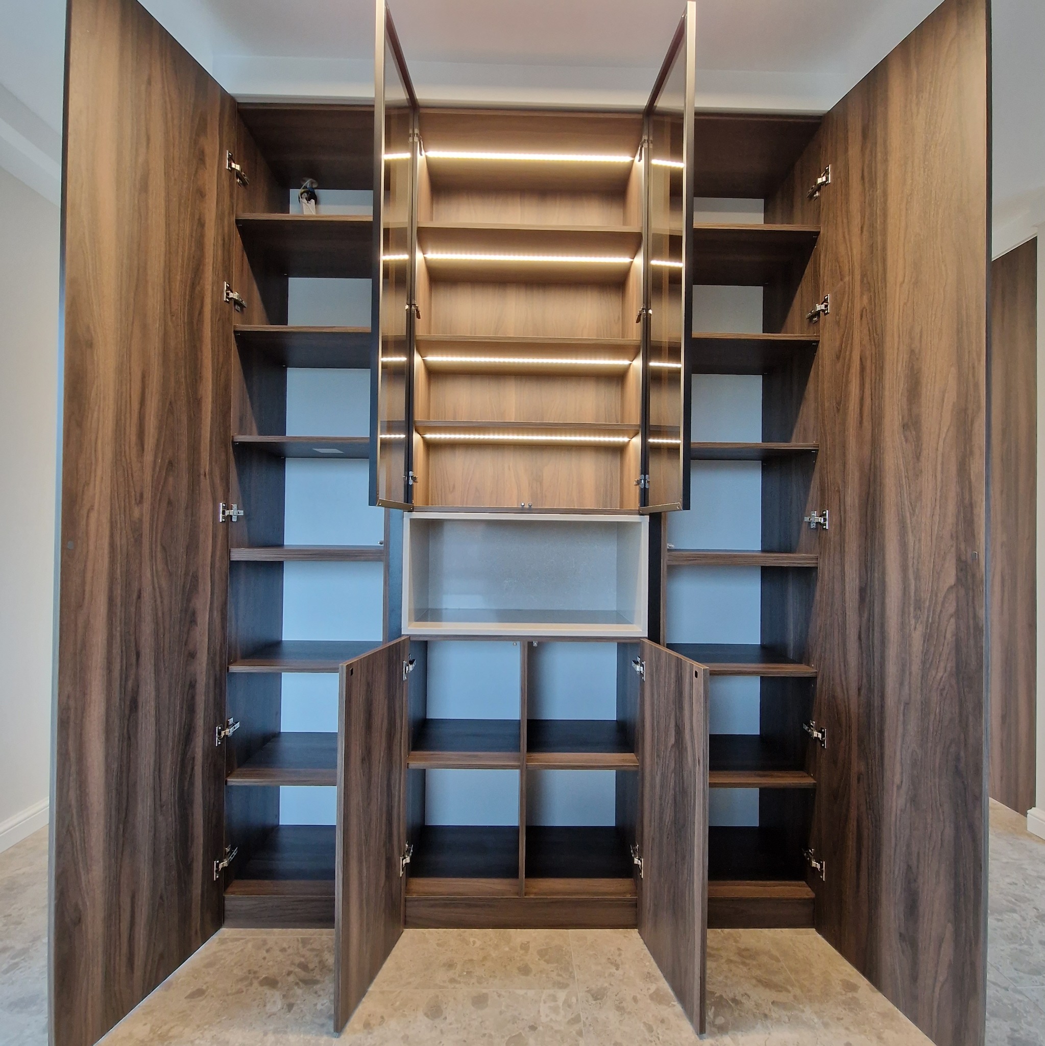 Wnętrze dużej, drewnianej szafy z otwartymi drzwiami, podświetlone listwami LED na półkach, widok od frontu na marmurową podłogę.