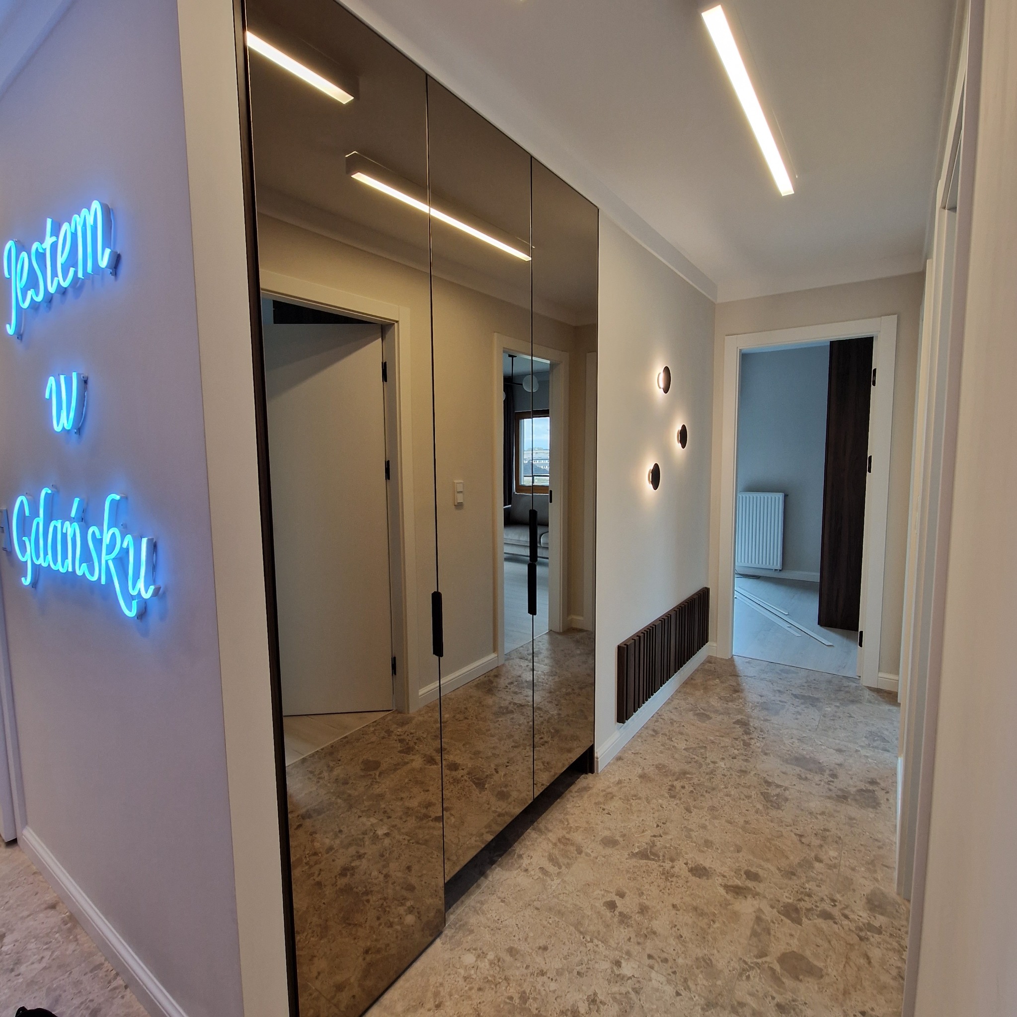Korytarz z lustrzaną szafą przesuwną, odbijającą wnętrze mieszkania, z neonem 'Jestem w Gdańsku' na ścianie.