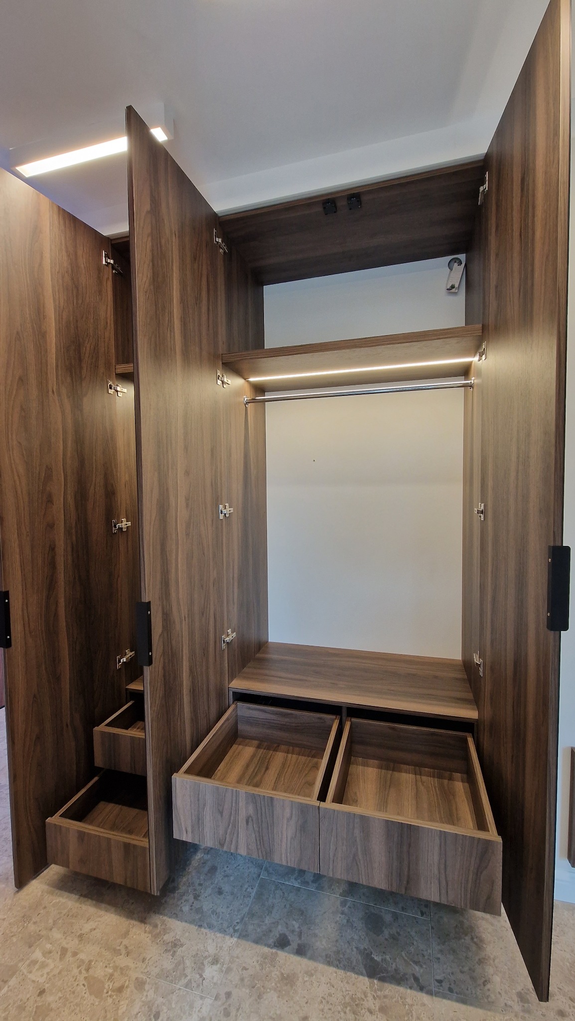 Otwarta szafa wnękowa z drewna orzechowego z szufladami i oświetleniem LED, widoczne zawiasy i uchwyty.