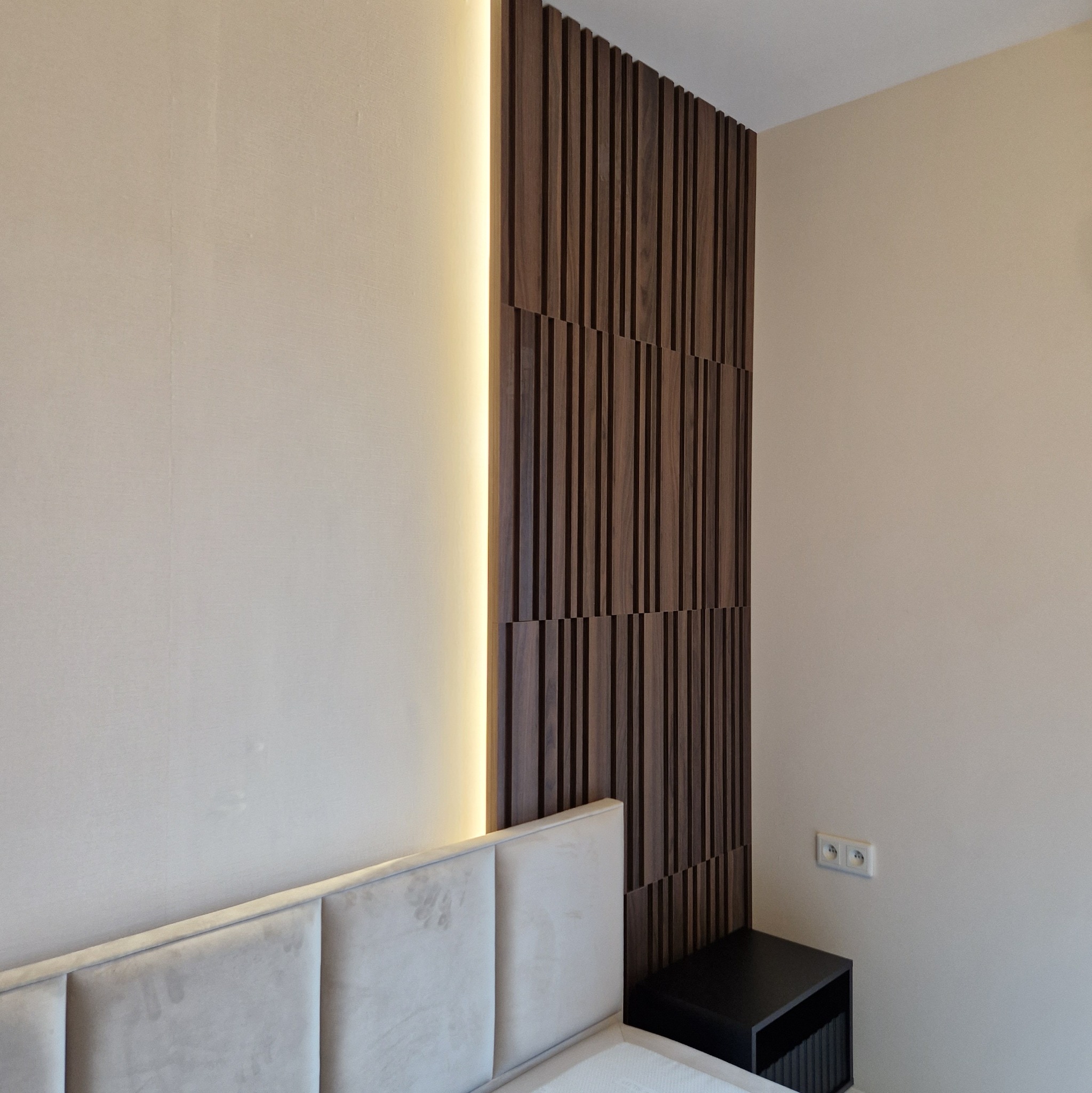 Panel ścienny z ciemnego drewna z podświetleniem LED, zamontowany za wezgłowiem łóżka i obok szafki nocnej, widoczne gniazdka elektryczne w ścianie.