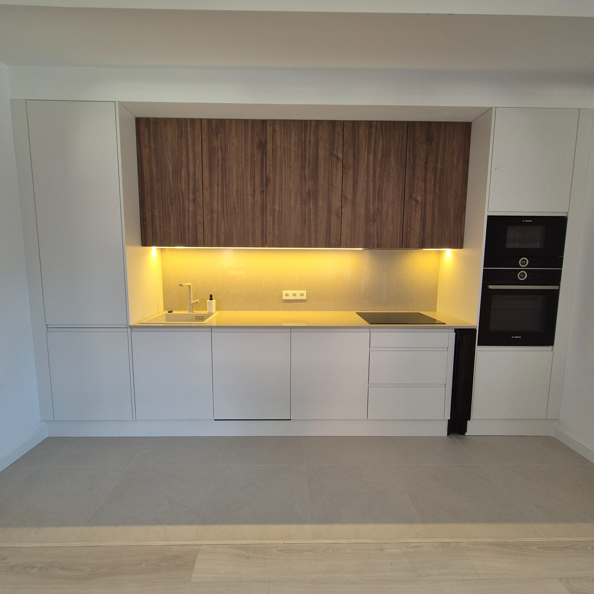 Nowoczesna kuchnia z białymi szafkami dolnymi i górnymi, uzupełniona drewnianymi szafkami nad blatem, zintegrowane oświetlenie LED pod szafkami podkreśla jasny blat i minimalistyczny zlew.