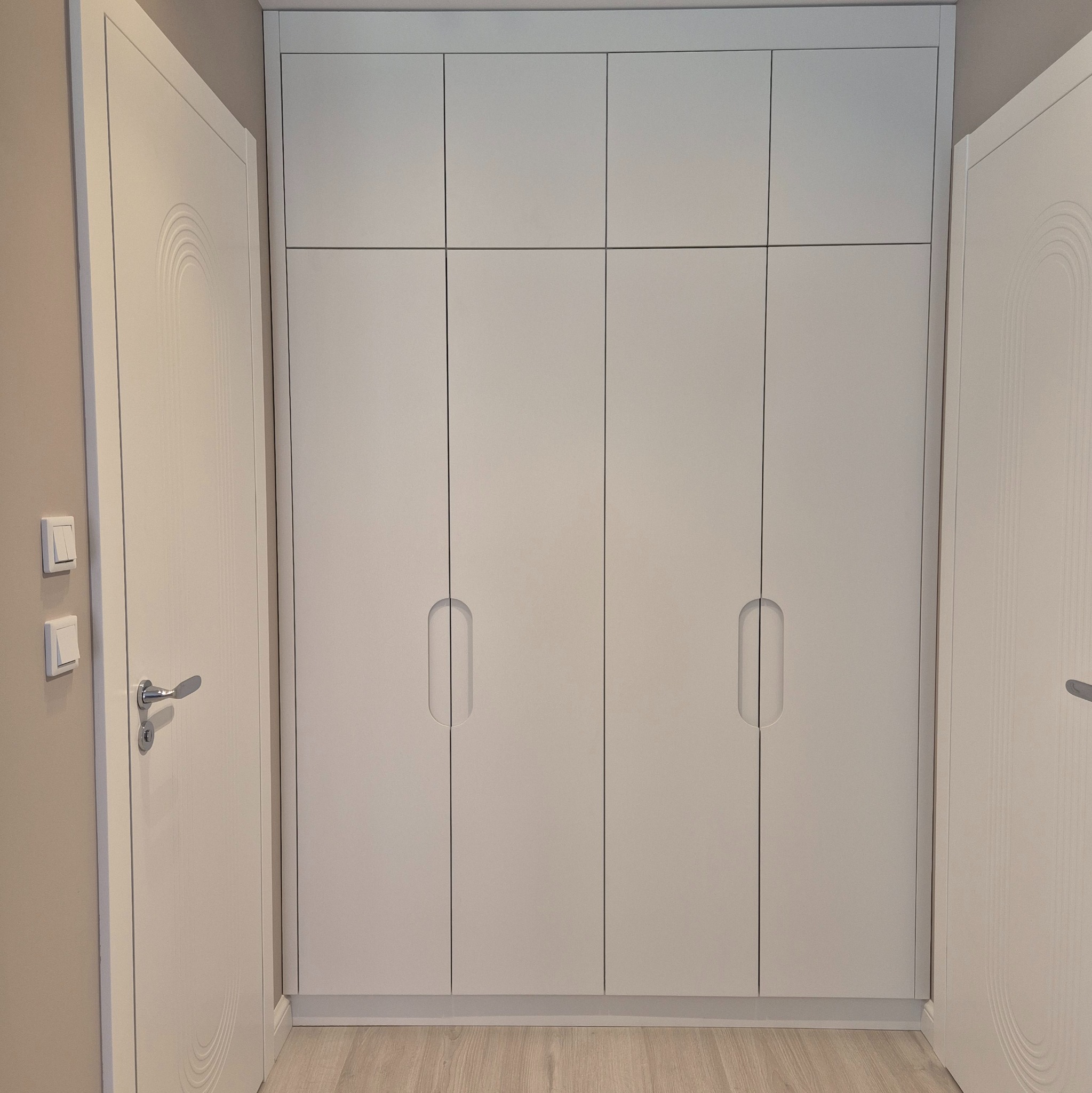 Biała szafa wnękowa z sześcioma drzwiami, podłoga z paneli, drzwi pokojowe z dekoracyjnym wzorem łuku.