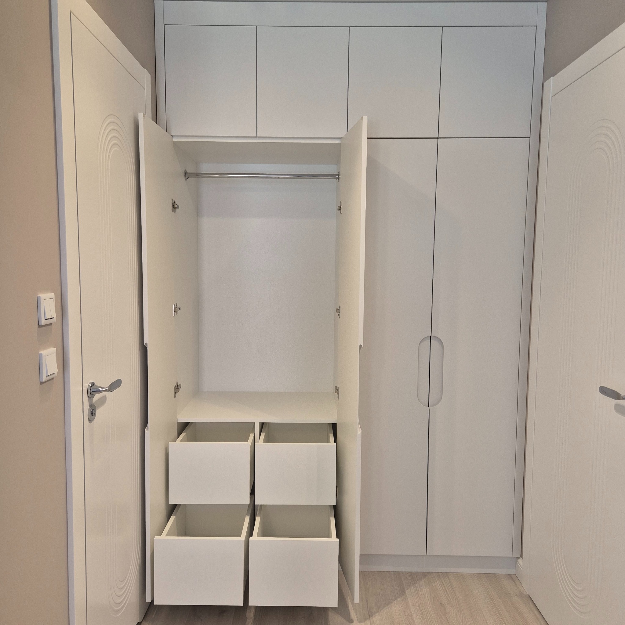 Biała szafa wnękowa z otwartymi drzwiami, widoczne półki i szuflady, minimalistyczny design, drzwi z subtelnym zdobieniem, jasne wnętrze.