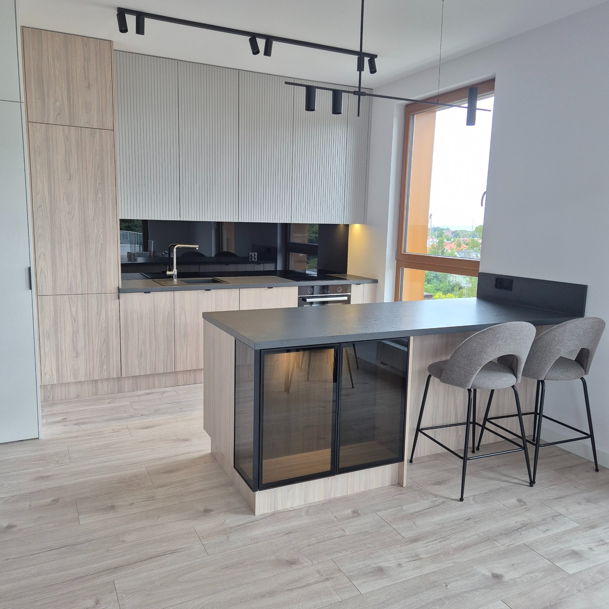 Nowoczesna kuchnia z drewnianymi szafkami, czarnym blatem i barkiem z szarymi hokerami, oświetlona reflektorami na szynoprzewodzie.