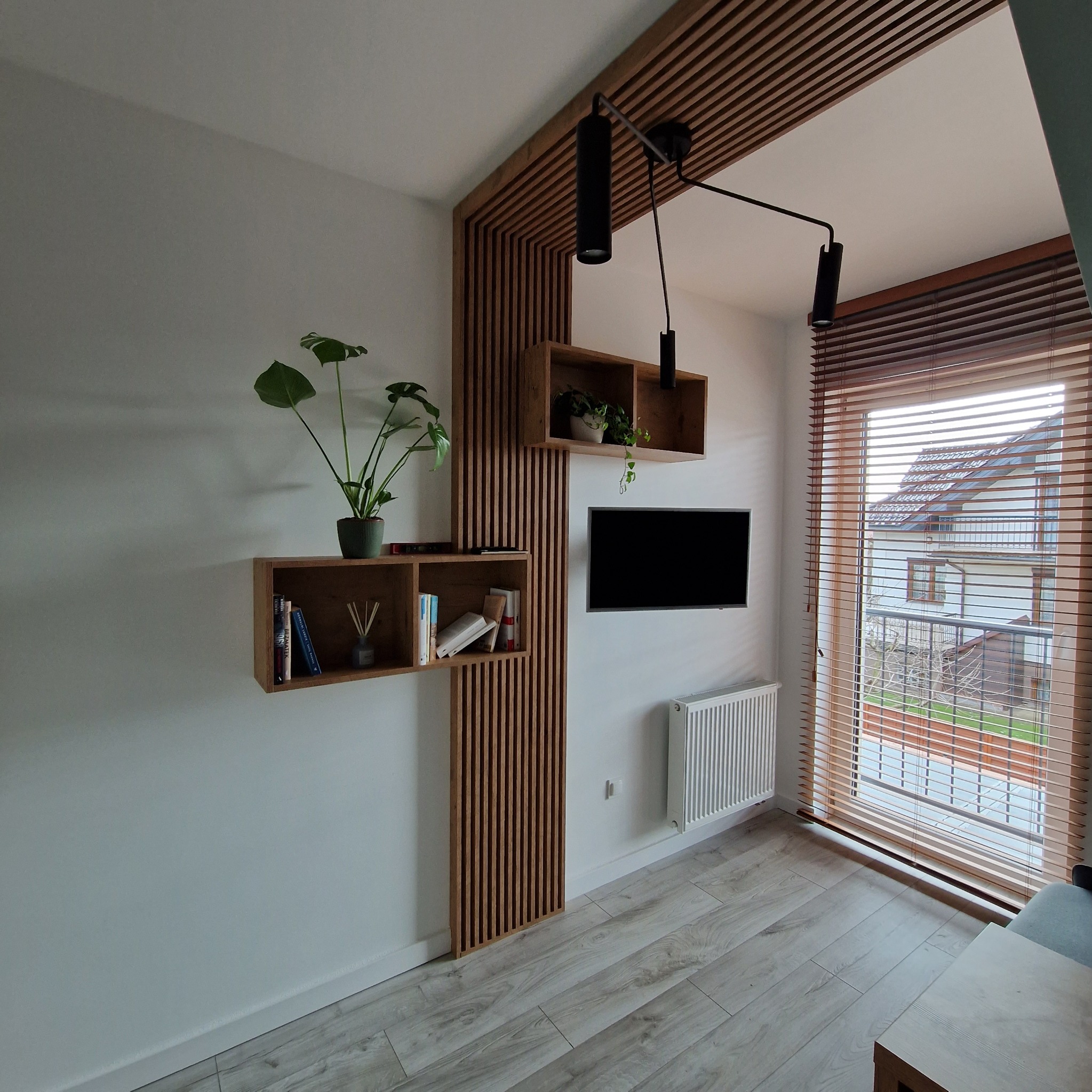 Nowoczesny salon z drewnianą zabudową ściany, półkami, roślinami, czarną lampą sufitową i telewizorem