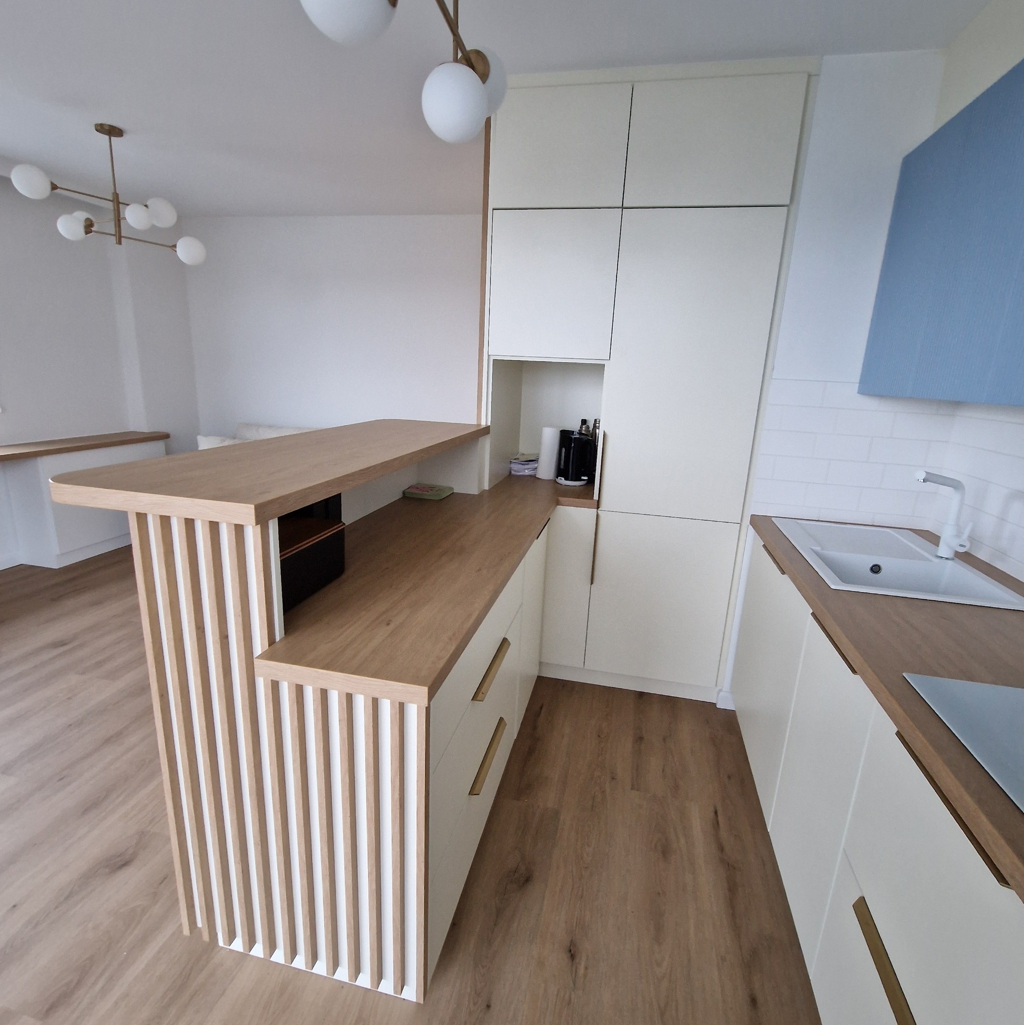 Jasna kuchnia z drewnianym blatem i wyspą z pionowymi listwami, nowoczesne oświetlenie i białe płytki na ścianie.