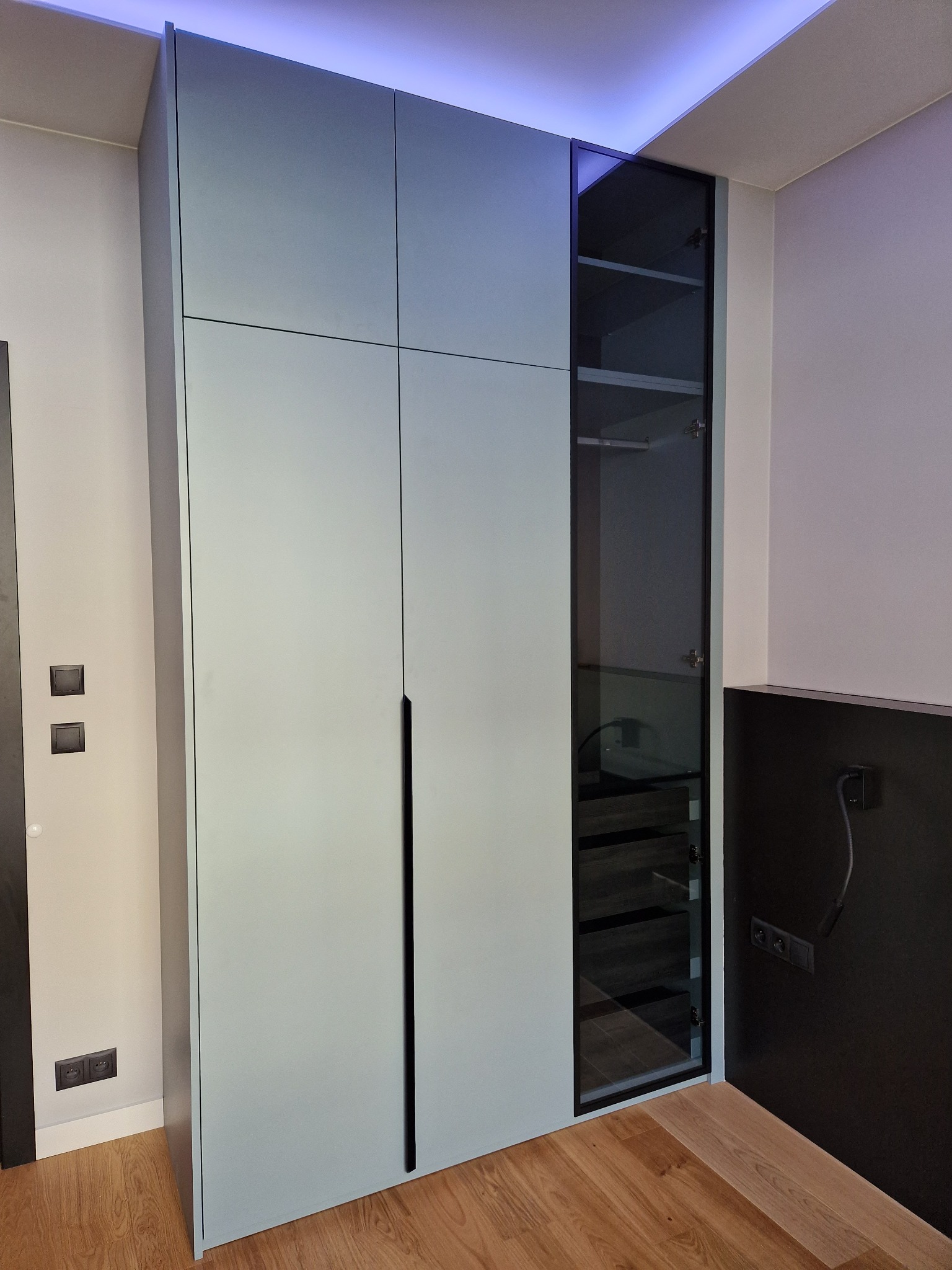 Pudrowo-niebieska szafa wnękowa z czarnymi, pionowymi uchwytami i przeszkloną witryną, podświetlona niebieskim światłem LED u góry, na tle drewnianej podłogi i ściany z czarnymi akcentami.