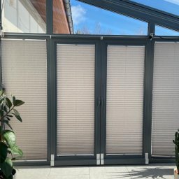 Bona Małgorzata Ptach - Beżowe plisy okienne zamontowane wewnątrz szklanej werandy, z widokiem na błękitne niebo, z roślinami doniczkowymi po bokach.