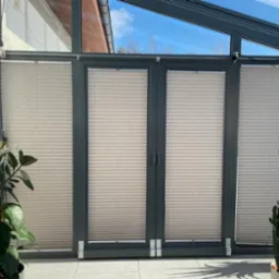 Beżowe plisy okienne zamontowane wewnątrz szklanej werandy, z widokiem na błękitne niebo, z roślinami doniczkowymi po bokach.