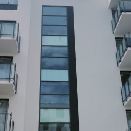 Bona Małgorzata Ptach - Fasada budynku z długim oknem w czarnej ramie, widoczne balkony z szklanymi balustradami, częściowo zakryte folią ochronną.