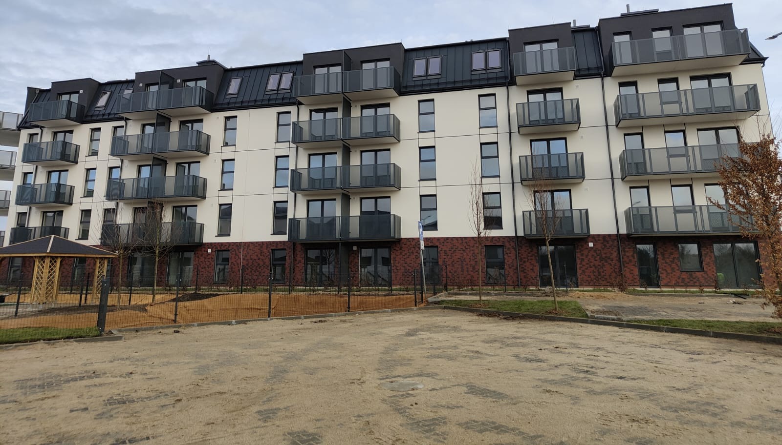 Nowoczesny apartamentowiec w Gdyni z oknami PCV i aluminiowymi balkonami z szklanymi balustradami, widok z poziomu gruntu na plac budowy.