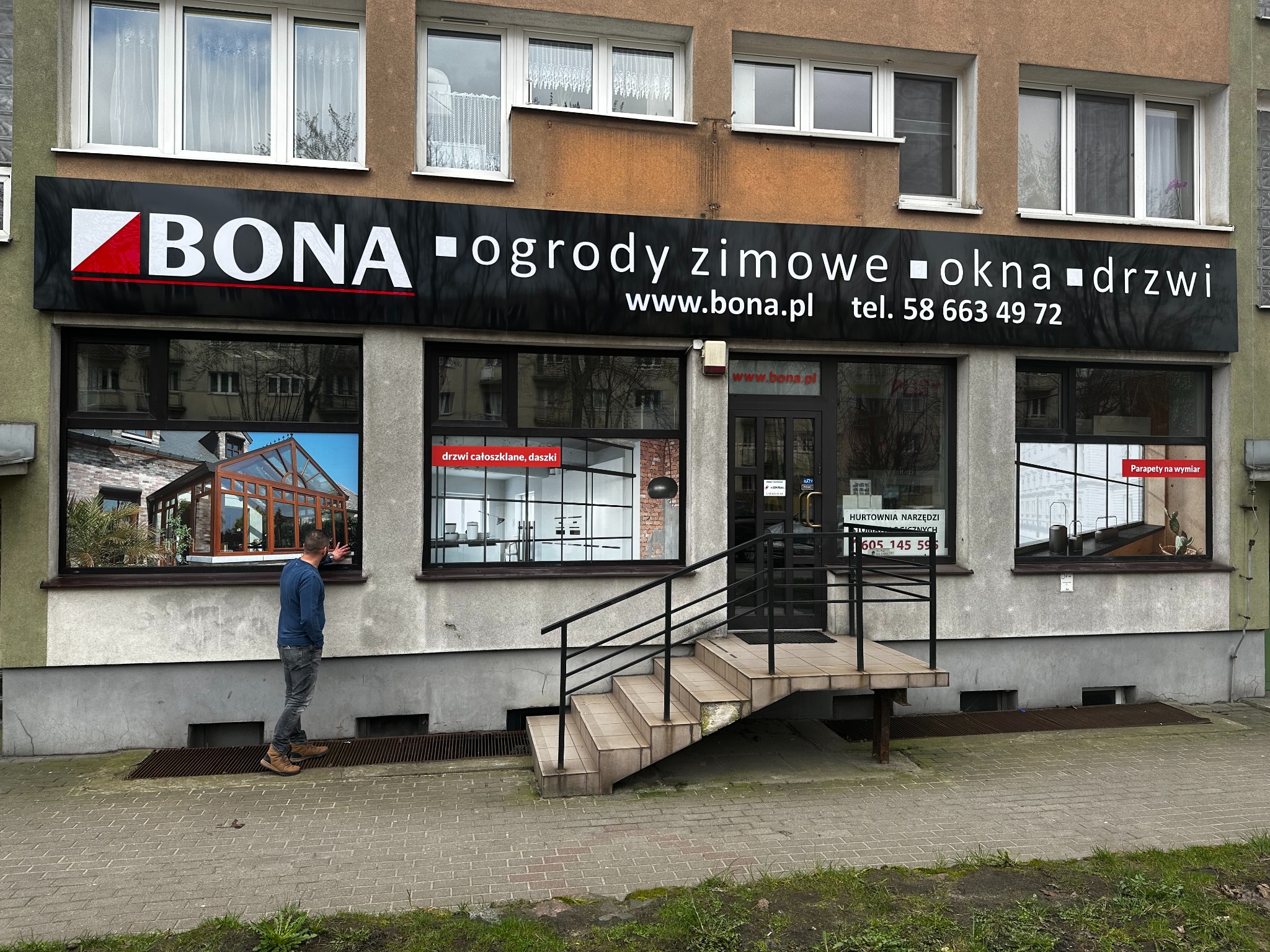 Fasada budynku z szyldem firmy 'BONA' oferującej ogrody zimowe, okna i drzwi. W witrynach wystawione przykładowe realizacje. Mężczyzna ogląda ekspozycję.