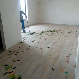 Integral Electrics and Constuction - Układanie płytek podłogowych: pracownik zamiata pozostałości po użyciu klipsów i klinów poziomujących, widoczne równe rzędy płytek imitujących drewno w jasnym odcieniu.