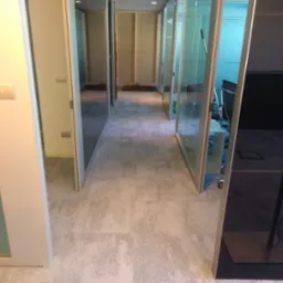 Korytarz biurowy z szarą podłogą imitującą beton, przeszklone ściany działowe z białymi ramami, widoczne gniazdka elektryczne na ścianie i kabel zasilający zwisający z sufitu.