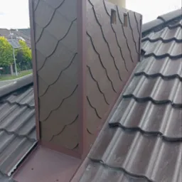 Obróbka komina z brązową blachą i łupkiem imitującym dachówki, na tle brązowej dachówki ceramicznej.