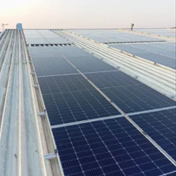 Rząd paneli słonecznych zamontowanych na blaszanym dachu, widoczny z góry pod kątem, elementy mocujące i komin wentylacyjny w tle.