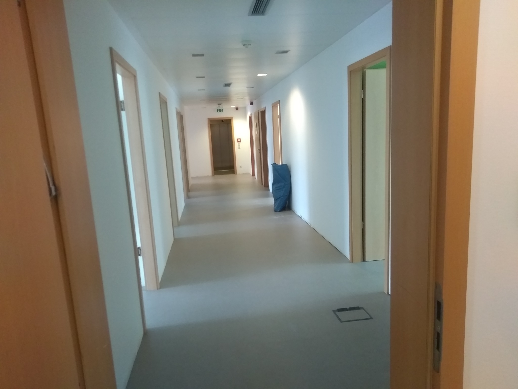 Długi korytarz w nowym budynku z białymi ścianami, drewnianymi drzwiami i szarą podłogą, z workiem budowlanym w tle i widoczną kratką wentylacyjną w podłodze.