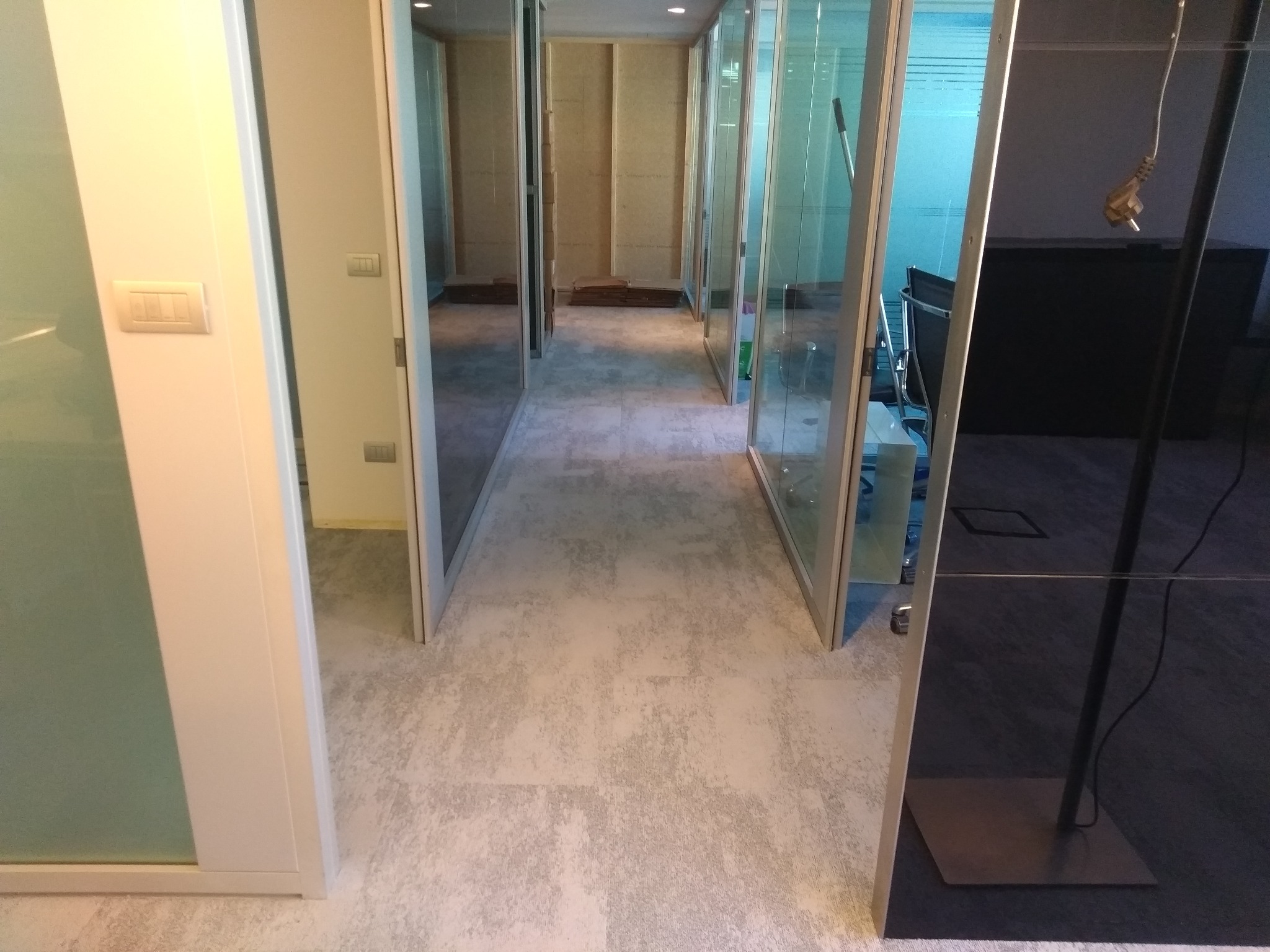 Korytarz biurowy z szarą podłogą imitującą beton, przeszklone ściany działowe z białymi ramami, widoczne gniazdka elektryczne na ścianie i kabel zasilający zwisający z sufitu.