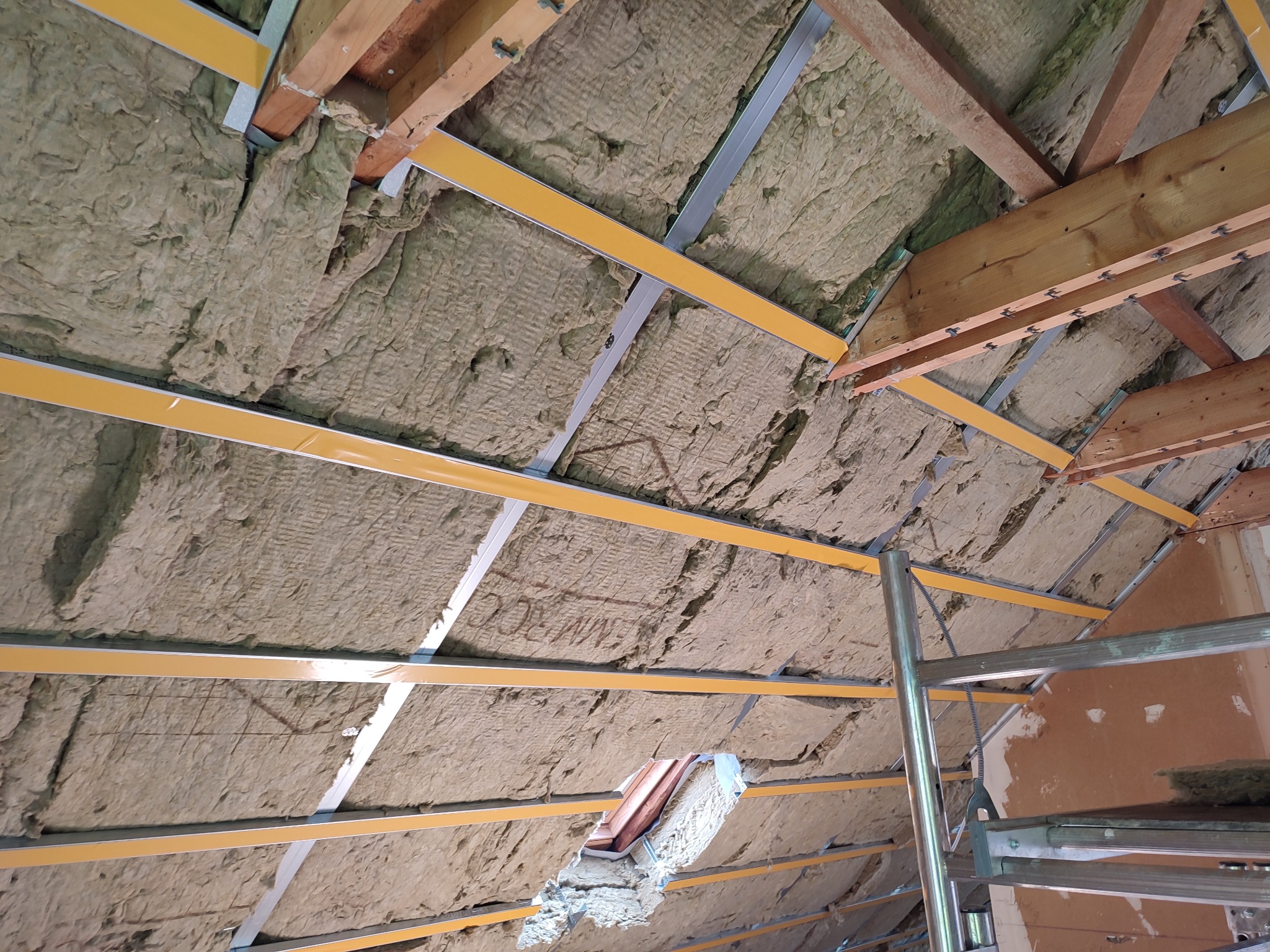Wnętrze poddasza w trakcie ocieplania wełną mineralną, widoczne drewniane krokwie, metalowe profile mocujące i fragment rusztowania.