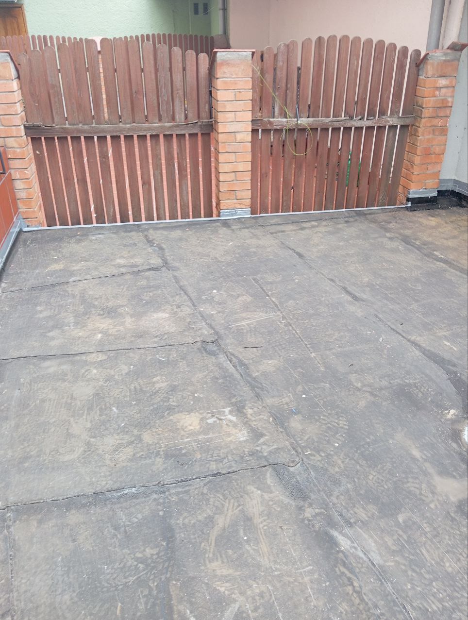 Płaski dach pokryty papą z widocznymi śladami stóp, w tle drewniany płot z ceglanymi słupkami.