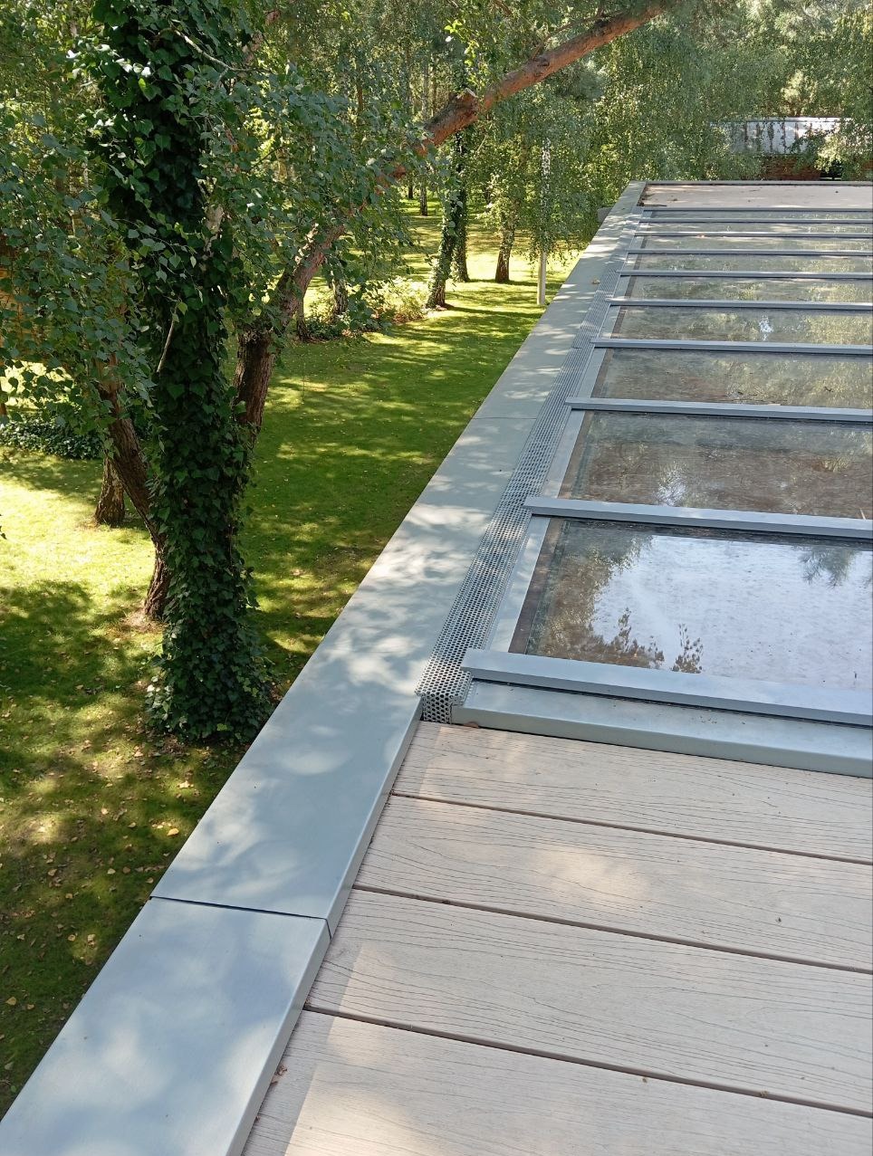 Szklany dach tarasu z drewnianą podłogą kompozytową i systemem odprowadzania wody, widok z góry na zewnątrz, z drzewami w tle.