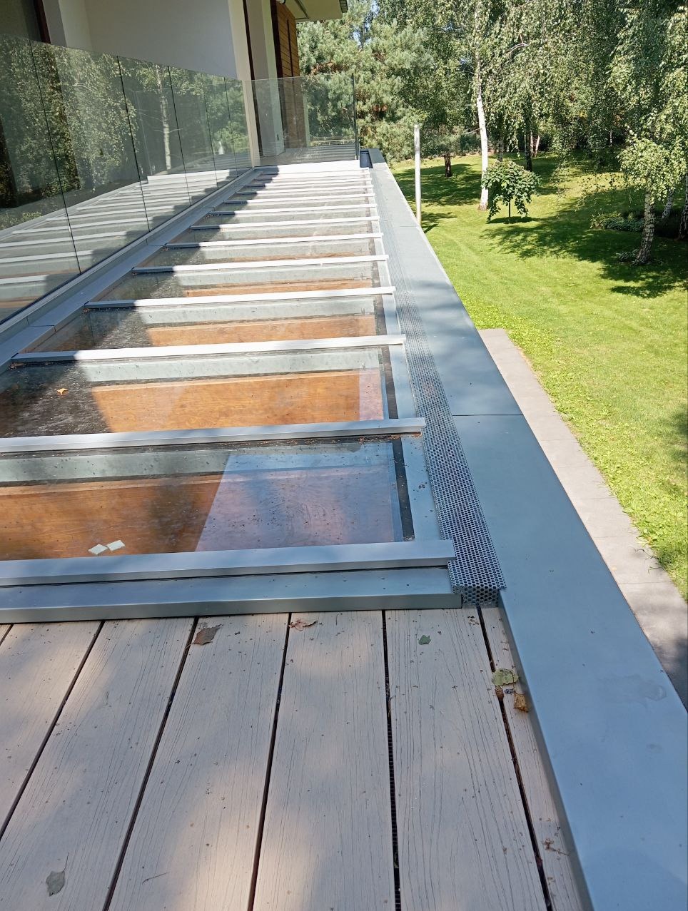 Zadaszony balkon z drewnianą podłogą i przeszklonym dachem, widok z zewnątrz na zieleń ogrodu.