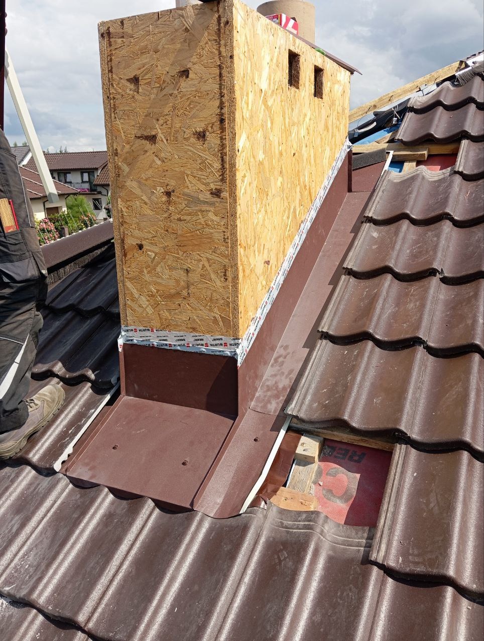 Montaż obudowy komina z płyty OSB na dachu z brązowej dachówki ceramicznej, widoczne okna w obudowie i elementy uszczelniające.