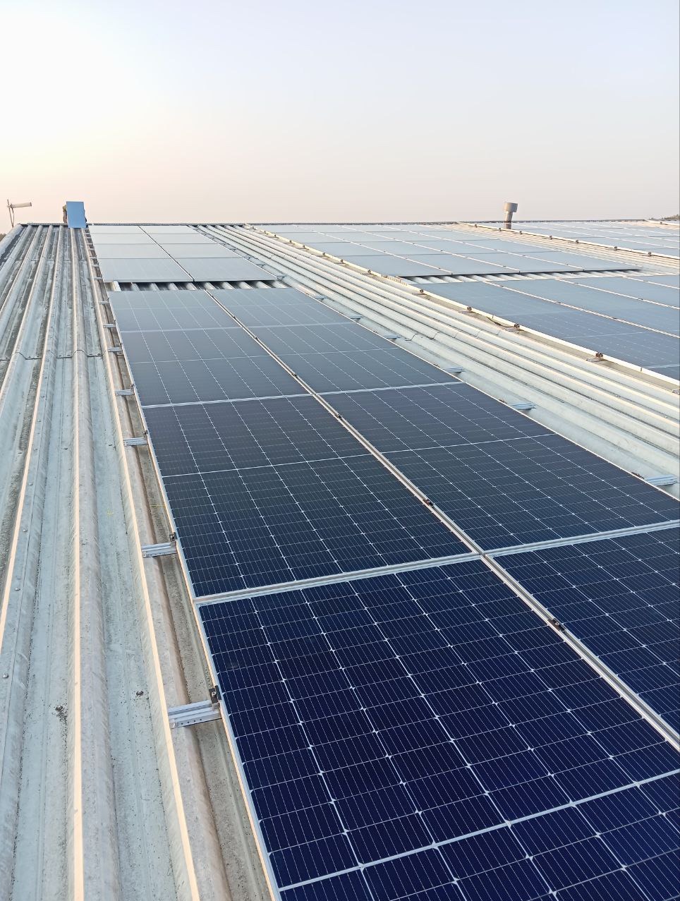 Rząd paneli słonecznych zamontowanych na falistym, metalowym dachu w pochmurny dzień, widoczne elementy montażowe i komin wentylacyjny w tle.