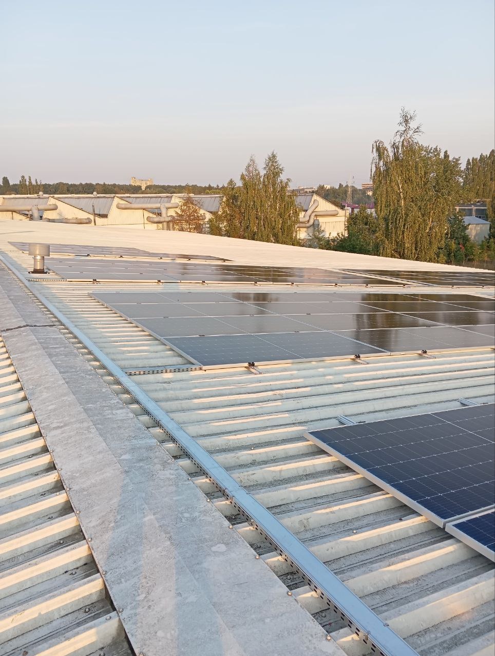 Instalacja paneli słonecznych na płaskim, metalowym dachu przemysłowym, widok z góry, z widocznymi elementami montażowymi i budynkami w tle.