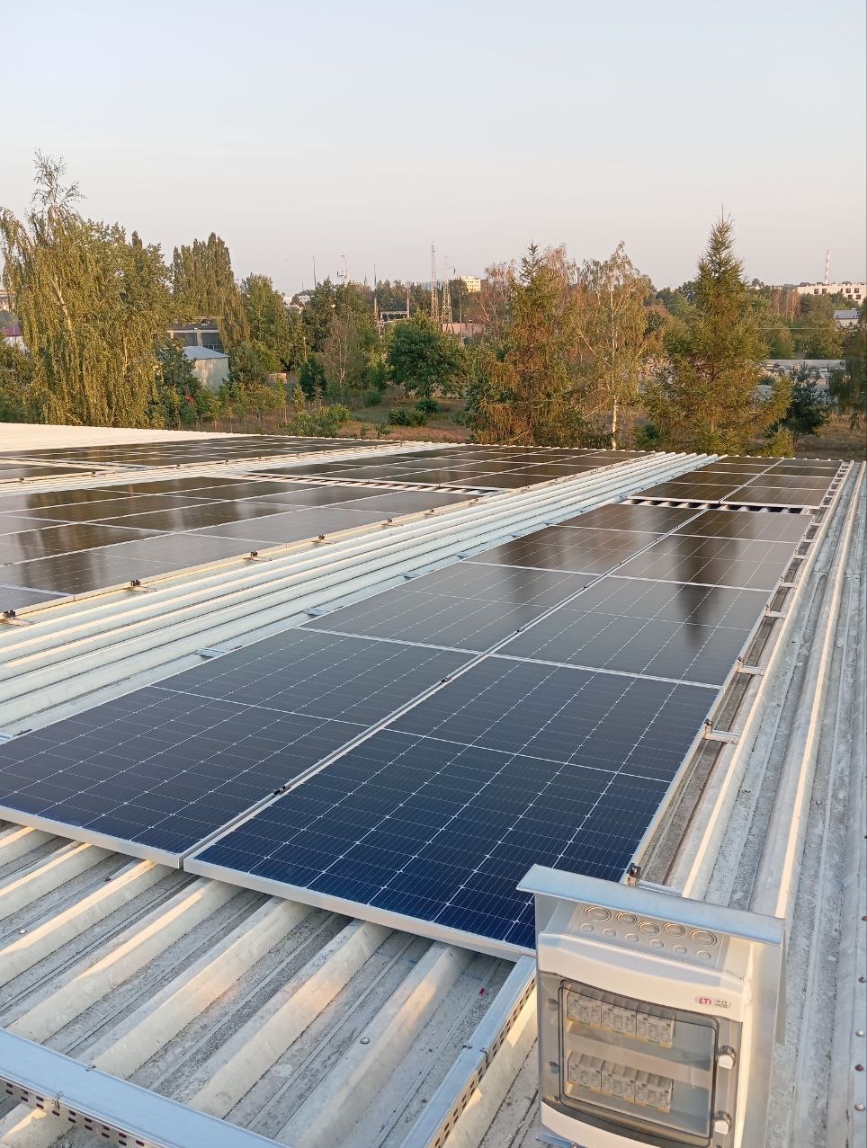 Instalacja paneli słonecznych na falistym dachu blaszanej konstrukcji, widoczna skrzynka z zabezpieczeniami elektrycznymi, zieleń drzew w tle.