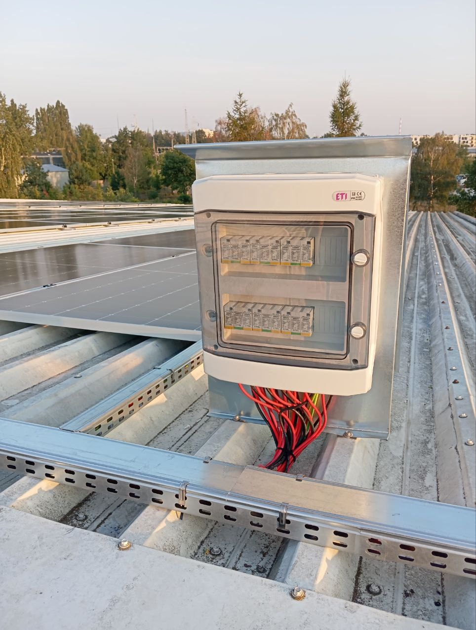 Rozdzielnica elektryczna z przewodami, zamontowana na dachu z panelami słonecznymi, widok z góry na instalację z metalowymi szynami montażowymi.