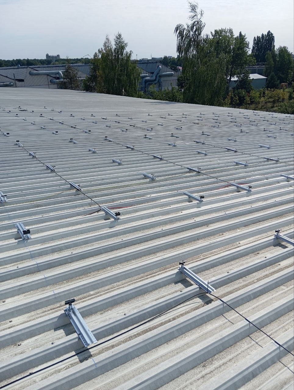 Montaż konstrukcji wsporczej pod panele słoneczne na dachu z blachy trapezowej, widoczne elementy mocujące i przewody elektryczne, w tle zieleń i budynki przemysłowe.